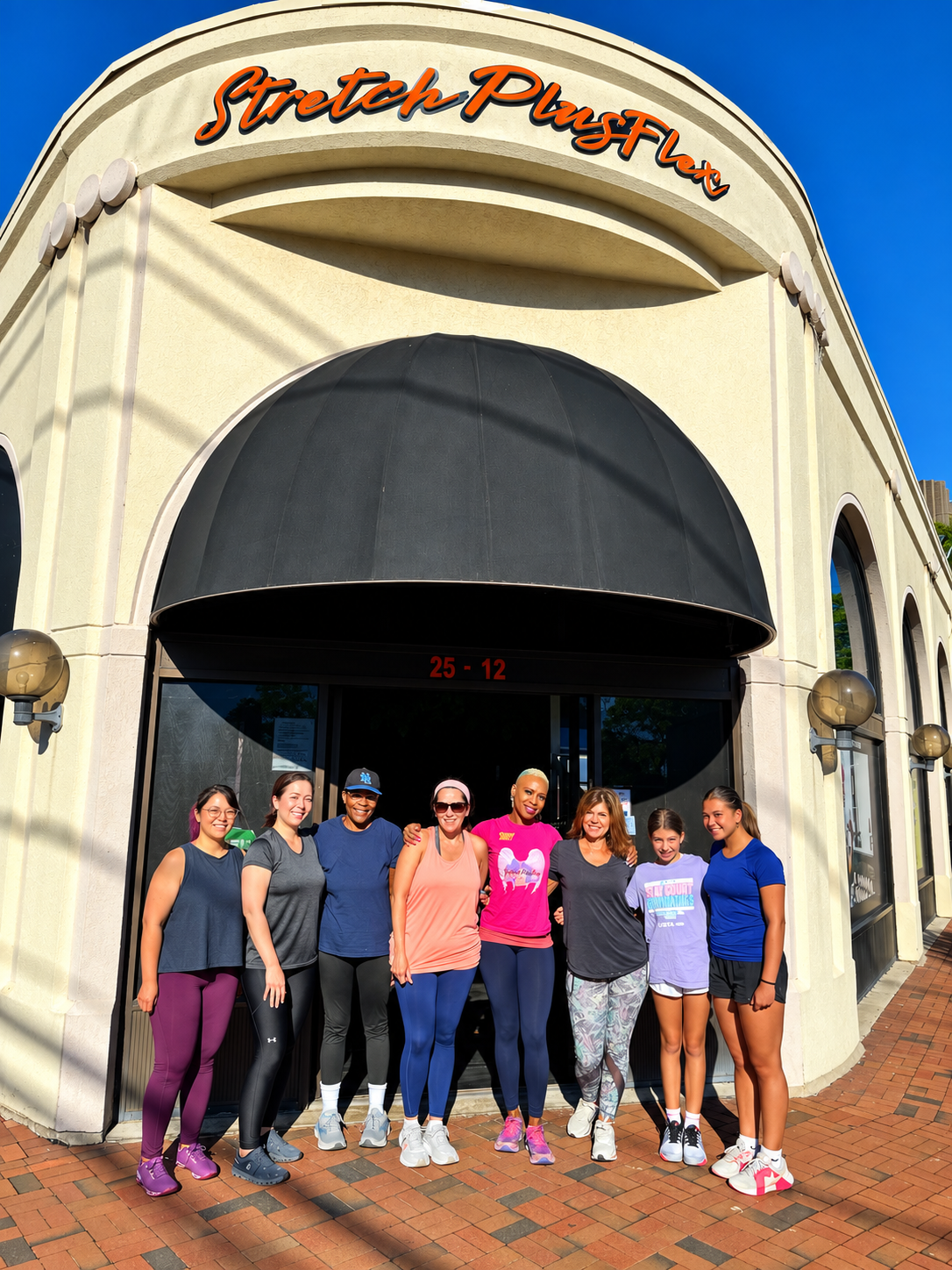 A group of eight women standing together outside the StretchPlusFlex studio in Great Neck, smiling and ready for a Women's Only class