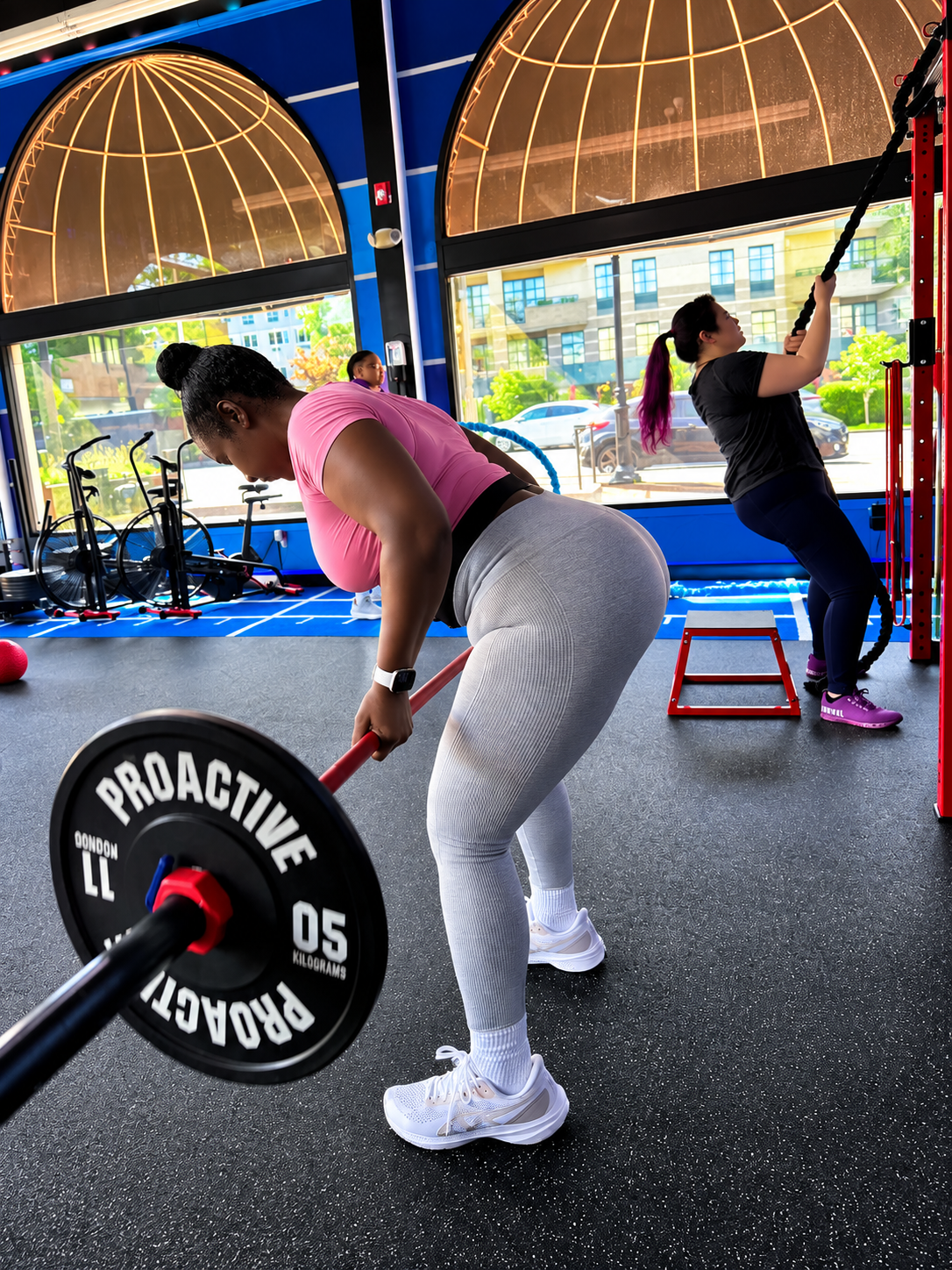 Member performing a barbell row in a small-group class at StretchPlusFlex, with another member on rope climbs in the background